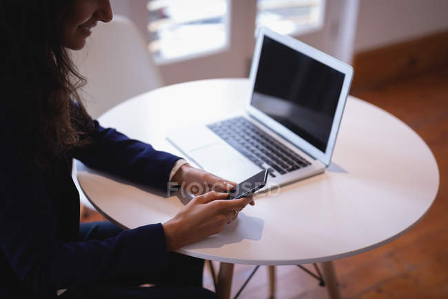 Side view of a brown hair mixed-race businesswoman using mobile phone while working on a laptop in the office — Stock Photo