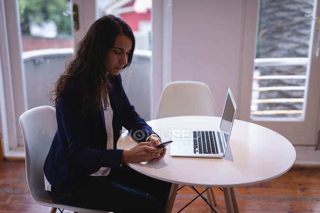 Seitenansicht einer schönen Frau mit braunem Haar, die ein Mobiltelefon benutzt, während sie im Büro an einem Laptop arbeitet — Stockfoto
