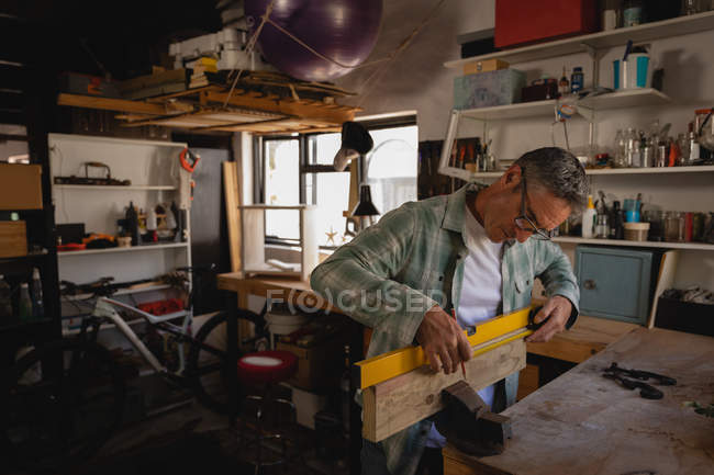 Vue latérale d'un charpentier mature caucasien attentif mesurant une planche de bois tout en travaillant en atelier — Photo de stock