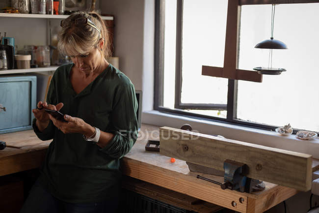 Frontansicht einer reifen kaukasischen Tischlerin, die ihr Handy benutzt, während sie sich in der Werkstatt gegen den Arbeitsplan lehnt — Stockfoto