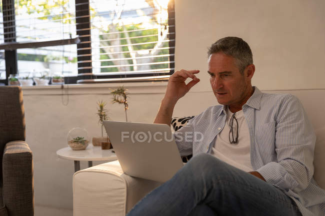 Front view of mature Caucasian man using laptop while sitting on sofa in living room at home — Stock Photo