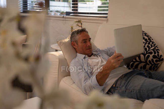 Vista lateral do homem caucasiano maduro usando laptop enquanto deitado no sofá na sala de estar em casa — Fotografia de Stock