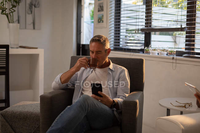 Vorderansicht eines reifen kaukasischen Mannes, der Kaffee trinkt, während er sein Mobiltelefon auf dem Sessel im heimischen Wohnzimmer benutzt — Stockfoto