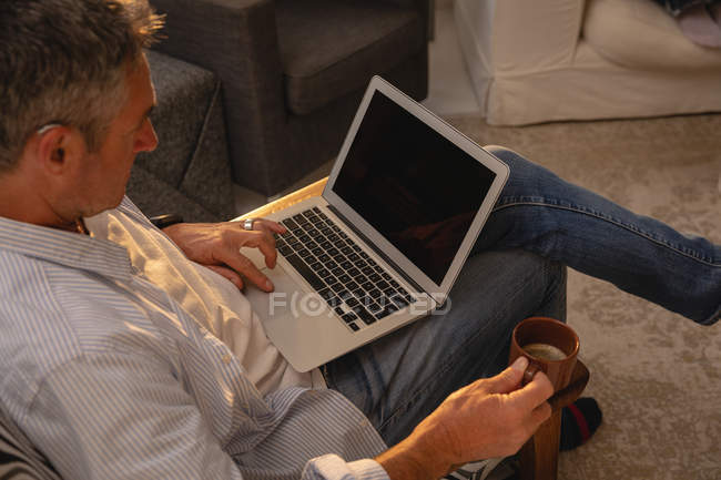 High angle view of mature Caucasian man holding coffee while using laptop in living room at home — Stock Photo