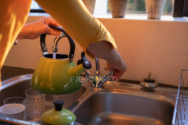 Sección media de la mujer que llena la tetera con agua en la cocina en casa - foto de stock