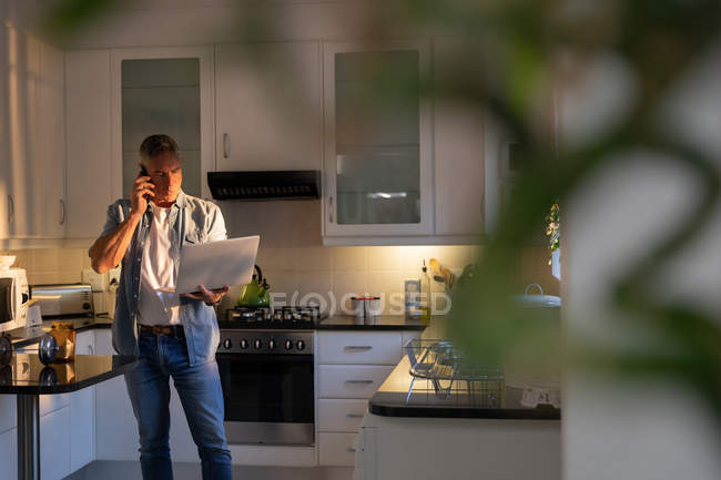 Вид спереди на взрослого кавказца, стоящего и разговаривающего по мобильному телефону, используя ноутбук с размытым растением на переднем плане на кухне дома — стоковое фото