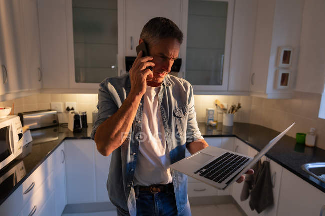 Вид спереду зрілого чоловіка, що стоїть і розмовляє на мобільному телефоні, використовуючи ноутбук на кухні вдома — стокове фото