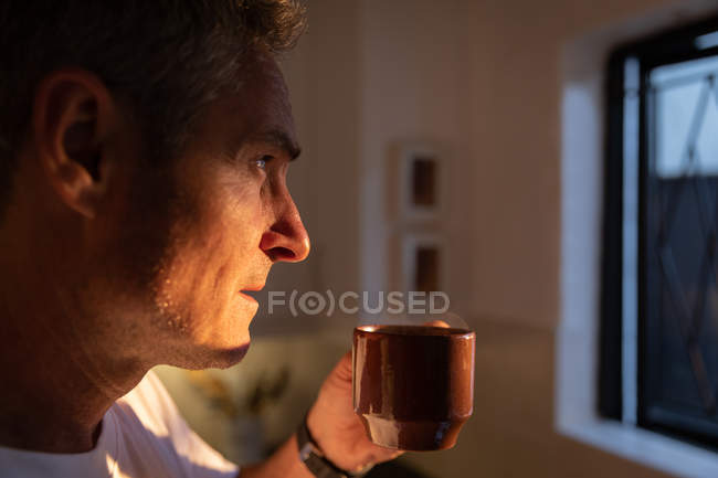 Vista lateral del hombre caucásico maduro reflexivo tomando café y mirando a través de la ventana en la cocina en casa al amanecer - foto de stock