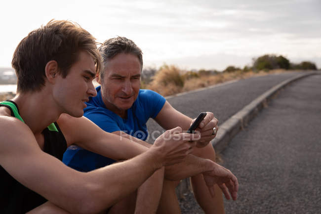 Vista lateral del padre y el hijo caucásicos mirando el teléfono móvil mientras se relaja en la carretera - foto de stock