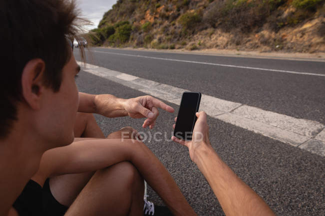 Por encima de la vista del hombro de padre e hijo mirando el teléfono móvil mientras se relaja en la carretera - foto de stock