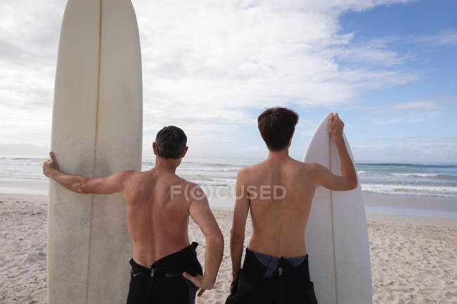 Rückansicht kaukasischer Vater und Sohn mit Surfbrett am Strand — Stockfoto