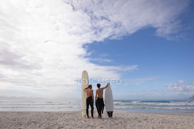 Vista trasera del padre y el hijo caucásicos de pie con tabla de surf en la playa - foto de stock