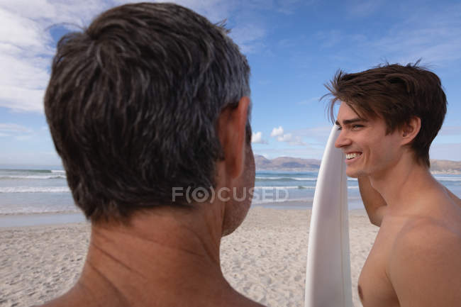 Rückansicht kaukasischer Vater und Sohn mit Surfbrett interagieren am Strand miteinander — Stockfoto