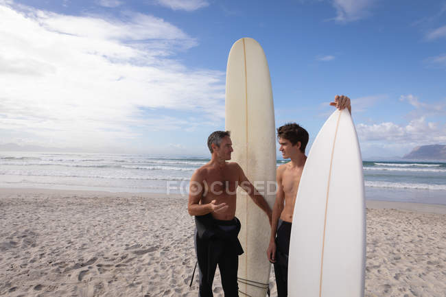 Vista frontal de pai e filho caucasiano com prancha interagem uns com os outros na praia — Fotografia de Stock