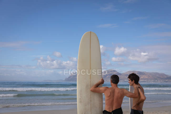 Rückansicht eines kaukasischen Vaters und Sohnes, die an einem sonnigen Tag mit Surfbrett am Strand stehen — Stockfoto