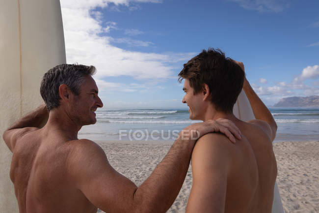 Vue arrière du père et du fils caucasiens avec planche de surf interagissent les uns avec les autres à la plage — Photo de stock