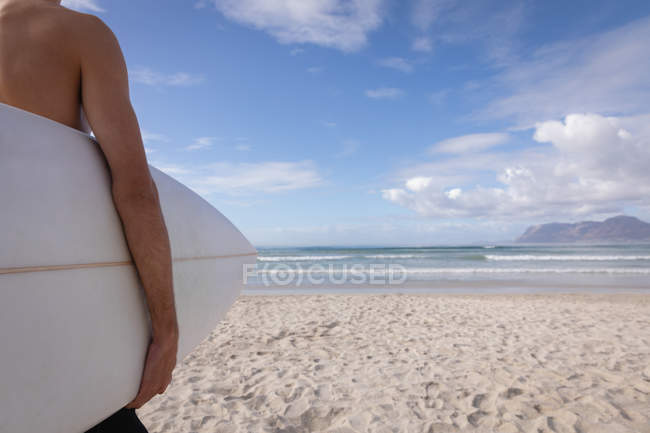 Mid-section of man standing with surfboard at beach on a sunny day — Stock Photo