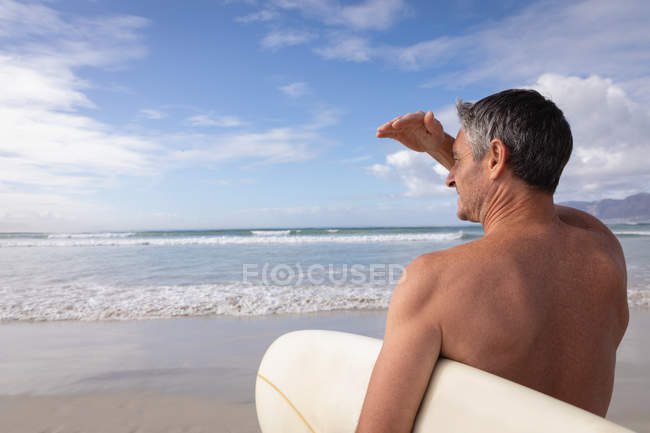Rückansicht eines kaukasischen Mannes, der an einem sonnigen Tag mit Surfbrett am Strand steht — Stockfoto
