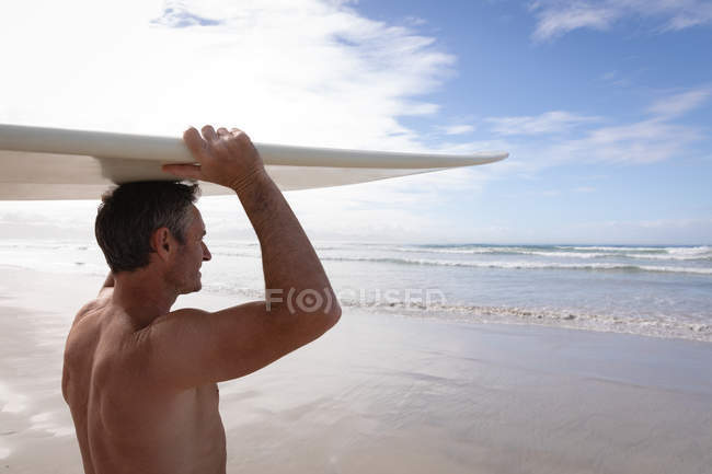 Боковой вид взрослого кавказца, стоящего с доской для серфинга на пляже в солнечный день — стоковое фото