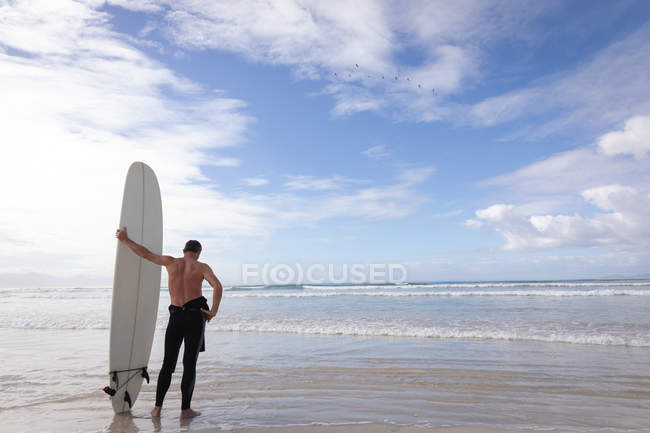 Vue arrière de l'homme caucasien debout avec planche de surf à la plage par une journée ensoleillée — Photo de stock