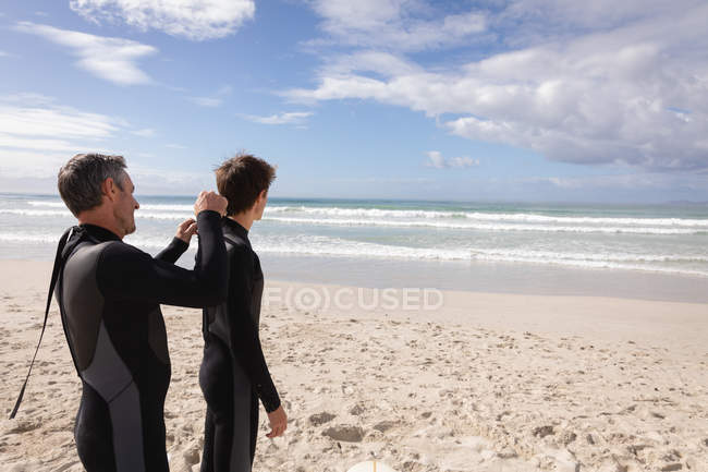 Rückansicht kaukasischer Vater hilft Sohn, an einem sonnigen Tag am Strand einen Neoprenanzug zu tragen — Stockfoto