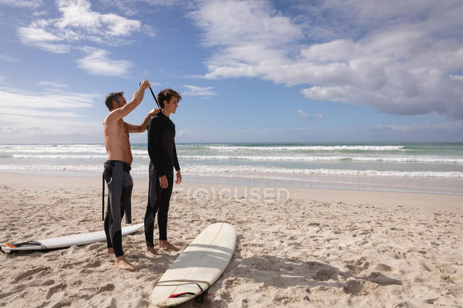 Vista lateral do pai caucasiano ajudar filho a usar roupa de mergulho na praia em um dia ensolarado — Fotografia de Stock