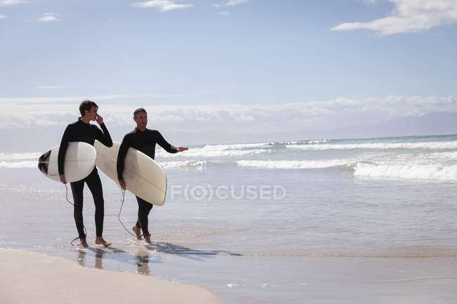 Side view of carefree Caucasian father and son with surfboard walking on beach — Stock Photo