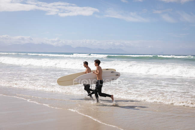 Vista lateral del padre y el hijo caucásicos con tabla de surf corriendo fuera del mar en el día soleado - foto de stock