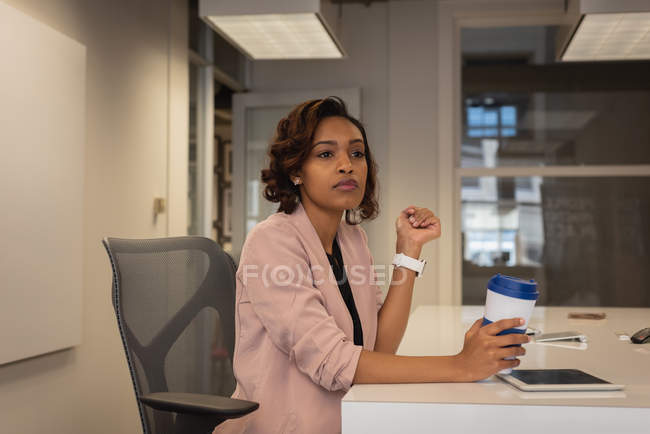 Vista frontal de la mujer de negocios reflexiva afroamericana con taza de café sentada en la silla en la oficina - foto de stock