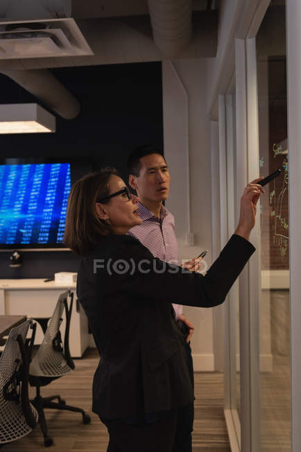 Vista lateral de la mujer de negocios caucásica mostrando carta a su colega asiático en la pared de vidrio en la oficina - foto de stock