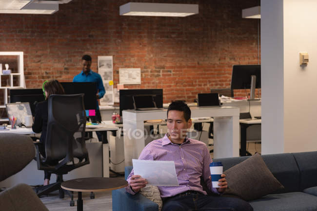 Vista frontal do empresário asiático olhando para o documento enquanto toma café no escritório — Fotografia de Stock