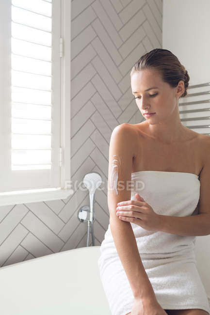 Belle femme appliquant lotion sur son corps dans la salle de bain à la maison — Photo de stock