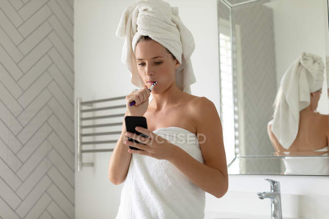 Красива жінка, використовуючи свій телефон, при чищенні зубів у ванній — стокове фото