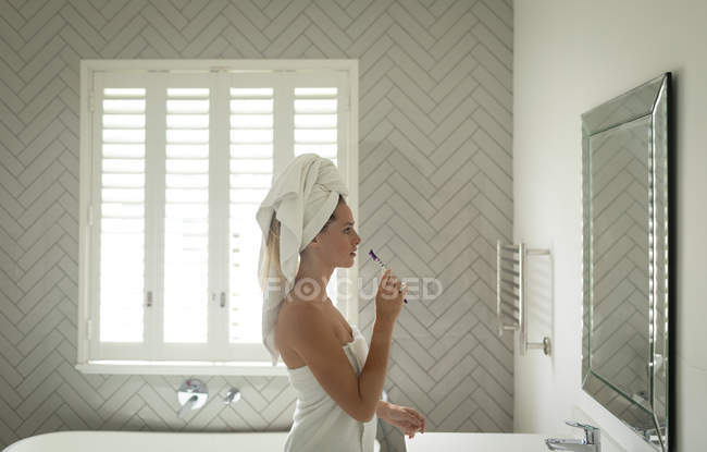 Боковой вид задумчивой красивой женщины, стоящей перед зеркалом, держа зубную щетку — стоковое фото