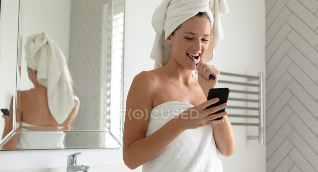 Красива жінка, використовуючи свій телефон, при чищенні зубів у ванній — стокове фото