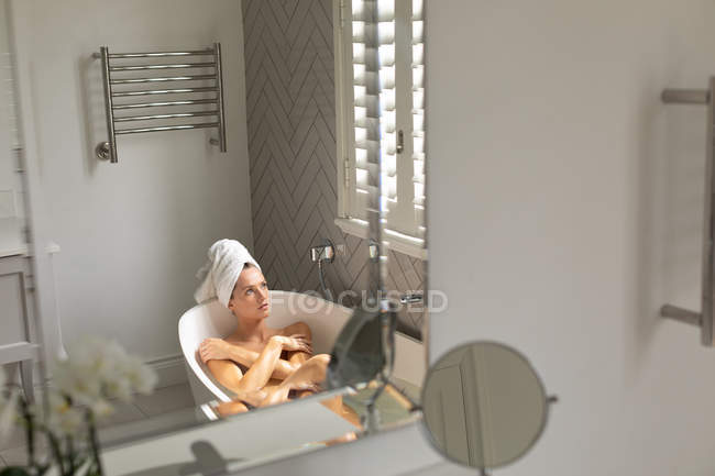 Nachdenkliche Frau, die in der Badewanne sitzt und zu Hause im Badezimmer aus dem Fenster schaut — Stockfoto