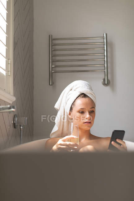 Mulher bonita tendo champanhe e verificando seu telefone na banheira em casa — Fotografia de Stock