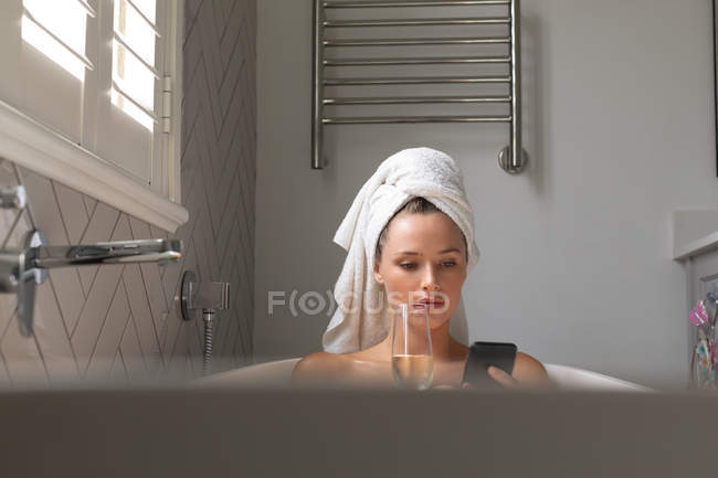 Красива жінка має шампанське і перевіряє свій телефон у ванній вдома — стокове фото