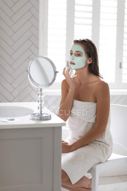 Жінка дивиться у дзеркало і наносить маску для обличчя після ванни у ванній — стокове фото