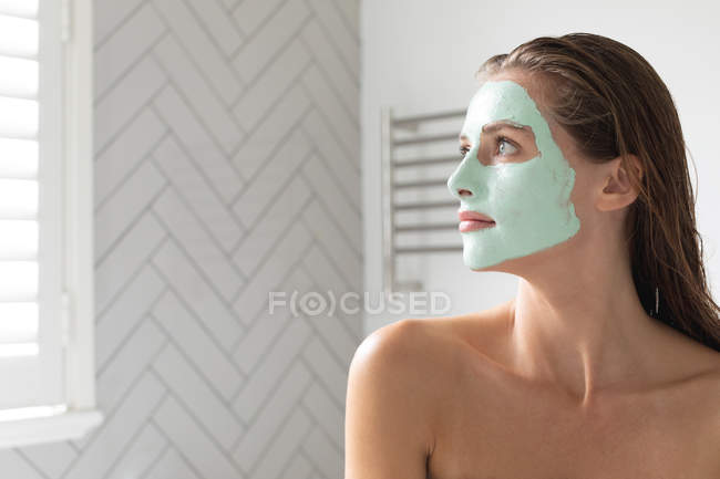 Nahaufnahme einer nachdenklichen Frau mit Gesichtsmaske, die im Badezimmer aus dem Fenster schaut — Stockfoto