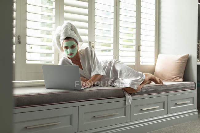Mulher de roupão deitada e usando um laptop perto da janela em casa — Fotografia de Stock