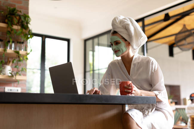 Mujer con taza de café utilizando un ordenador portátil en la cocina después de tomar un baño en la mañana - foto de stock