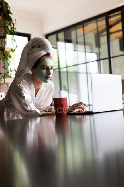 Hermosa mujer en albornoz utilizando el ordenador portátil en el mostrador de la cocina en casa - foto de stock