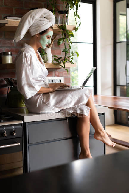 Mulher de roupão sentado no balcão da cozinha e usando laptop pela manhã — Fotografia de Stock