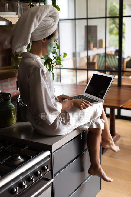 Mujer en albornoz sentado en el mostrador de la cocina y el uso de ordenador portátil por la mañana - foto de stock