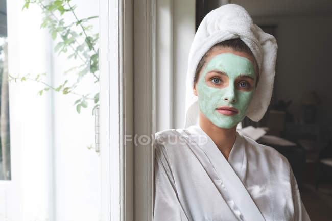 Beautiful woman in bathrobe wearing facial mask, leaning against the window — Stock Photo