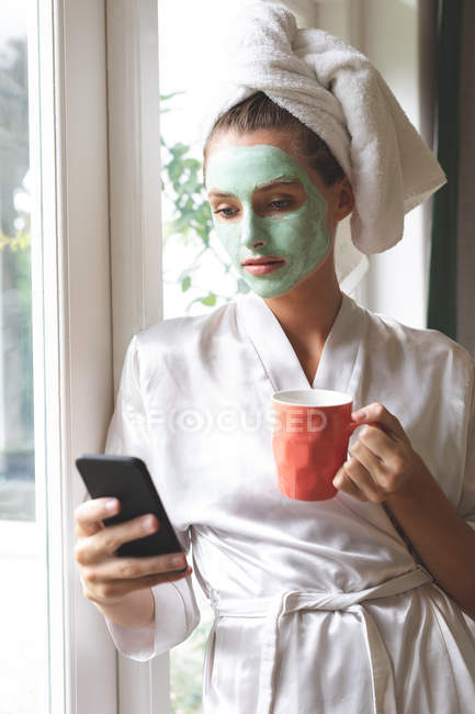 Красива жінка в масці для обличчя, використовуючи мобільний телефон біля вікна вдома — стокове фото