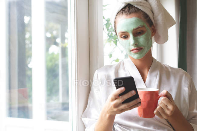 Mulher bonita em máscara facial usando telefone celular perto da janela em casa — Fotografia de Stock