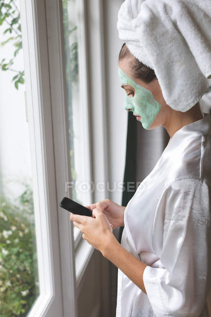 Schöne Frau mit Gesichtsmaske mit Handy in der Nähe des Fensters zu Hause — Stockfoto