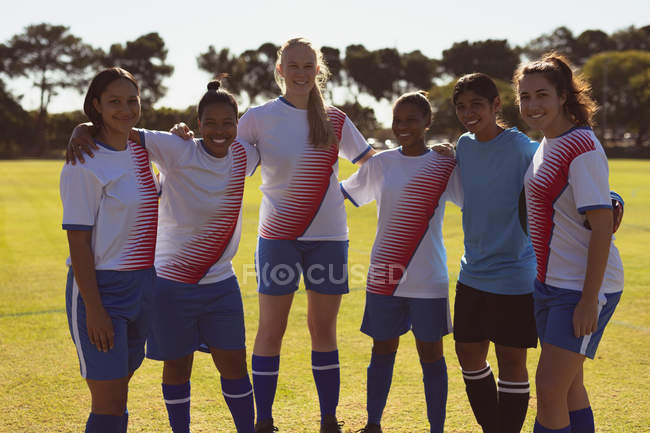 Портрет различных женщин-футболистов, стоящих обнимая друг друга на спортивной площадке в солнечный день — стоковое фото